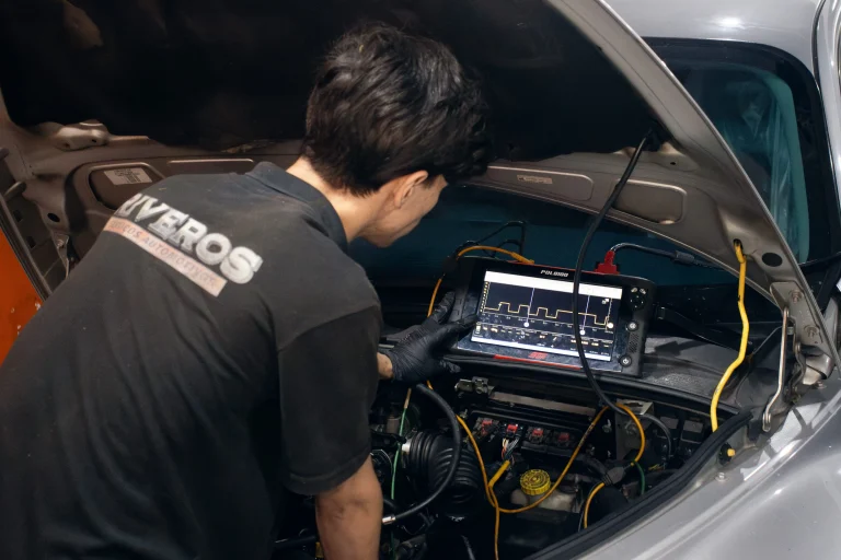 Mecanico vestindo uniforme preto escrito riveros oficina automotiva, fazendo diagnostico com osciloscópio em um veículo prata