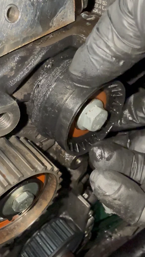 Mãos de um mecânico usando luvas pretas segurando um rolamento tensor de correia de motor. A peça está extremamente gasta, coberta de uma pasta escura de borra e resíduos de borracha derretida.